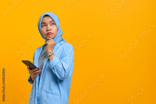 Pensive young Asian woman using mobile phone with fingers on chin over yellow background