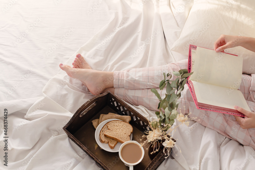 Woman reading a book and breakfast on dark wooden vintage tray in bed with light beige sheet and pillows. Flat lay, top view. Slow morning concept.