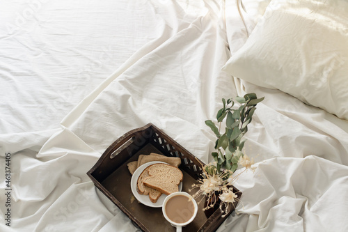 Breakfast on dark wooden vintage tray in bed with light beige sheet and pillows. Flat lay, top view. Slow romantic morning concept.