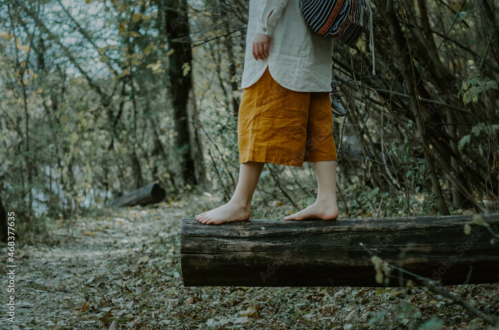 Person in saffron linen shorts and white shirt with backpack walking and balancing on a fallen tree trunk, in front of a forest path by a lake Person in saffron linen shorts and white shirt with backpack walking and balancing on a fallen tree trunk, in front of a forest path by a lake
