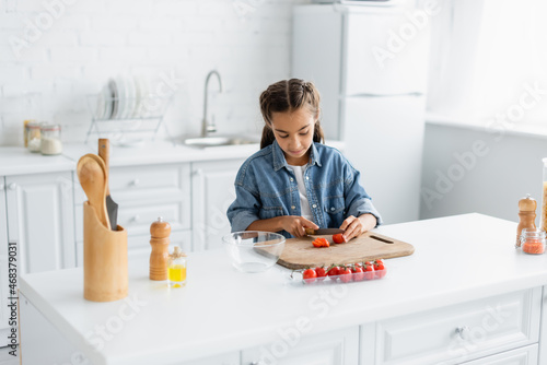 Preteen girl cutting cherry tomato near olive oil and bowl in kitchen