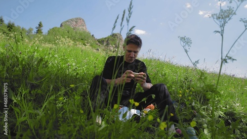 A young man is sitting on a slope and playing a game on his phone in a mountainous area. Tourism and travel