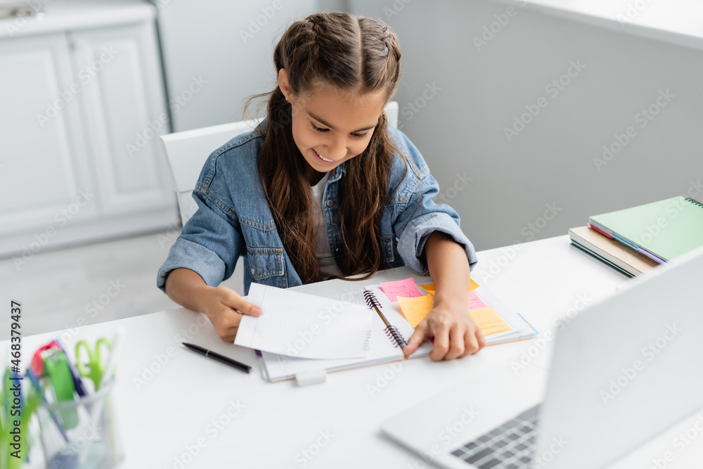 Happy kid tearing paper near notebook with sticky notes and blurred laptop at home Happy kid tearing paper near notebook with sticky notes and blurred laptop at home