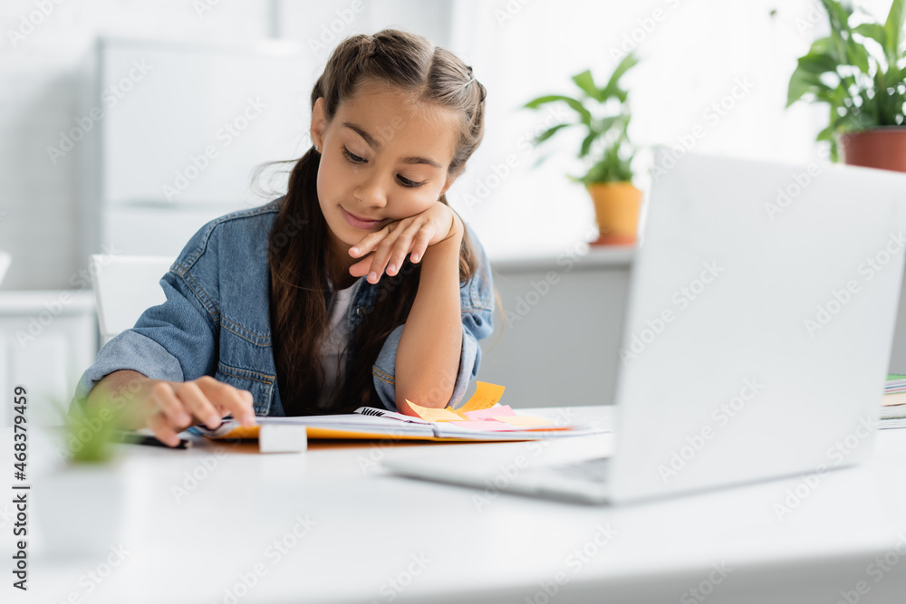 Kid looking at copy book near laptop during homework in kitchen Kid looking at copy book near laptop during homework in kitchen