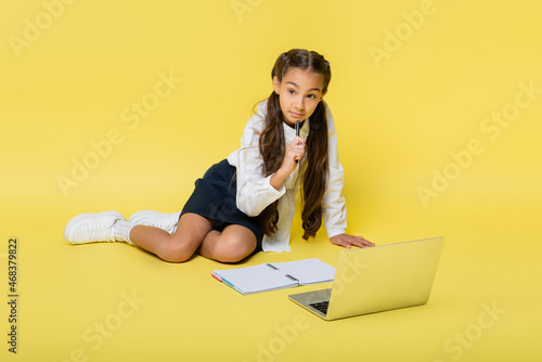 Pensive schoolkid holding pen near laptop and copy book on yellow background