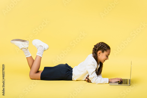 Side view of positive schoolchild using laptop while lying on yellow background