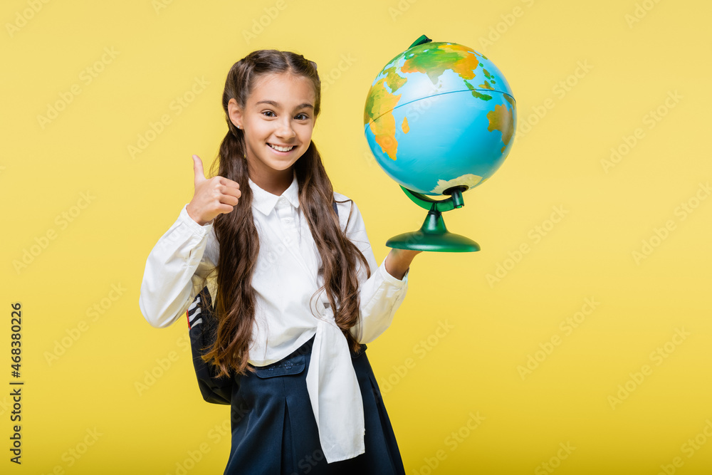 Fototapeta premium Smiling schoolgirl showing like and holding globe isolated on yellow