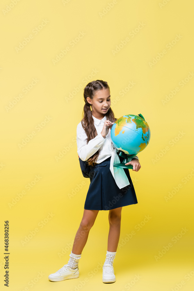 Positive schoolchild with backpack looking at globe on yellow background