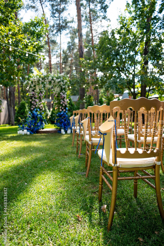 Outdoor wedding ceremony on a green lawn. Arch for a wedding ceremony of fresh flowers in white and blue and chairs in gold for guests. Soft selective focus.