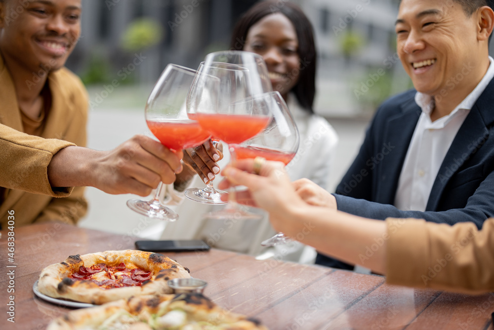 Multiracial business team toasting with wine while celebrating at outdoor cafe. Concept of teambuilding and corporate event. Idea of rest and leisure on job. People sitting at table with pizza