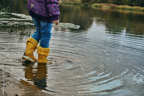 A child in yellow rubber boots stands in the water, blue jeans and a purple jacket, oversinking the weather. In rubber sabogs in water