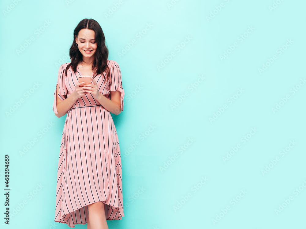 Young beautiful smiling female in trendy summer dress. Sexy carefree woman posing near blue wall in studio. Positive brunette model looking at cellphone screen. Holding smartphone and using apps Young beautiful smiling female in trendy summer dress. Sexy carefree woman posing near blue wall in studio. Positive brunette model looking at cellphone screen. Holding smartphone and using apps
