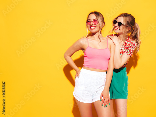 Two young beautiful smiling blond hipster female in trendy summer clothes. Sexy carefree women posing near yellow wall in studio. Positive models having fun. Cheerful and happy. In sunglasses