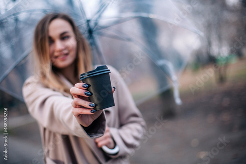 A young, beautiful girl with a disposable cup of coffee walks in the park under a transparent umbrella in the rain © Denis