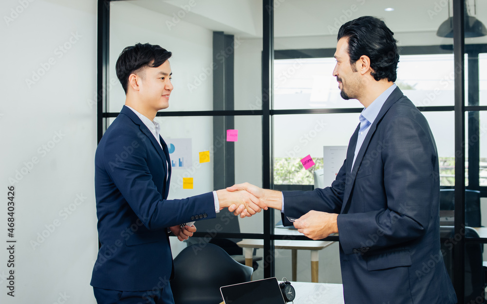 Two diverse handsome partner customer businessmen wearing formal suit, shaking hand to deal business, introduce themselves, exchange name card, smiling with success and confidence, standing in office.