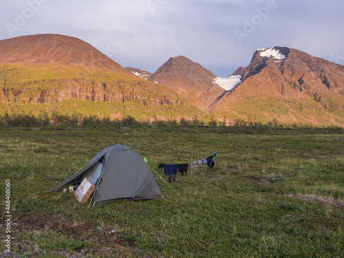 Green trekking tent with hanging clothes in beautiful nature with Akka mountain massif with snow  glacier and birch tree in Swedish Lapland landscape at Padjelantaleden hiking trail. Golden hour light
