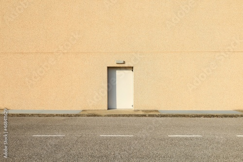 Fototapeta Naklejka Na Ścianę i Meble -  White door with a lamp above on yellow wall made of concrete mixed with small pebbles. Sidewalk and asphalt road in front. Background for copy space. 