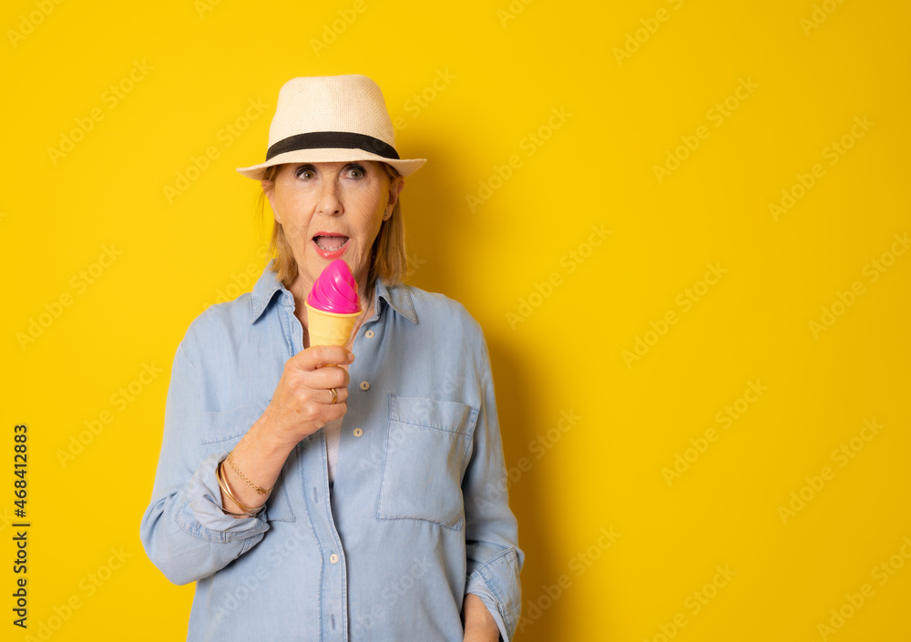 Senior woman in straw hat holding a cornet ice cream over isolated yellow background Senior woman in straw hat holding a cornet ice cream over isolated yellow background