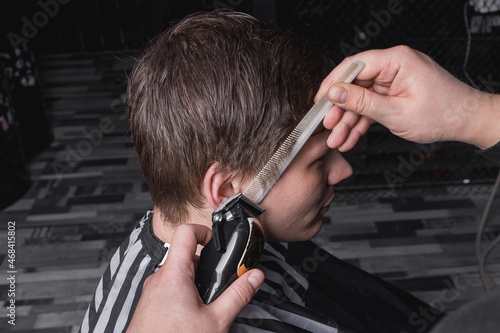 The hands of a barber or hairdresser cut the client's temple with dark hair with a automatic machine and a comb. Hairdressing
