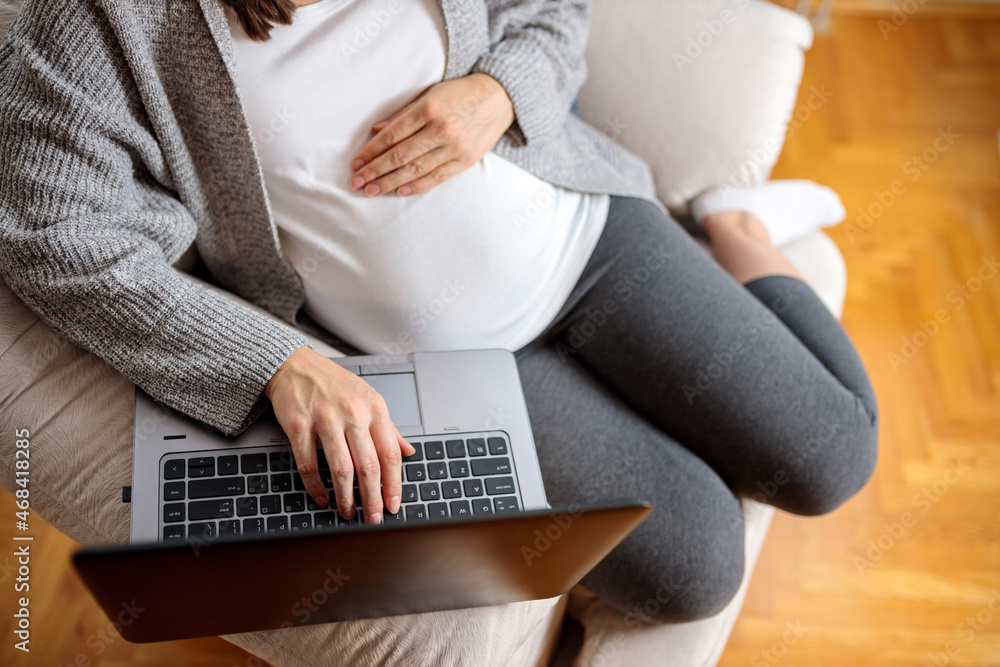 Naklejka premium High angle view of unrecognizable pregnant young woman sitting on a sofa and browsing internet on laptop. Pregnancy at home concept