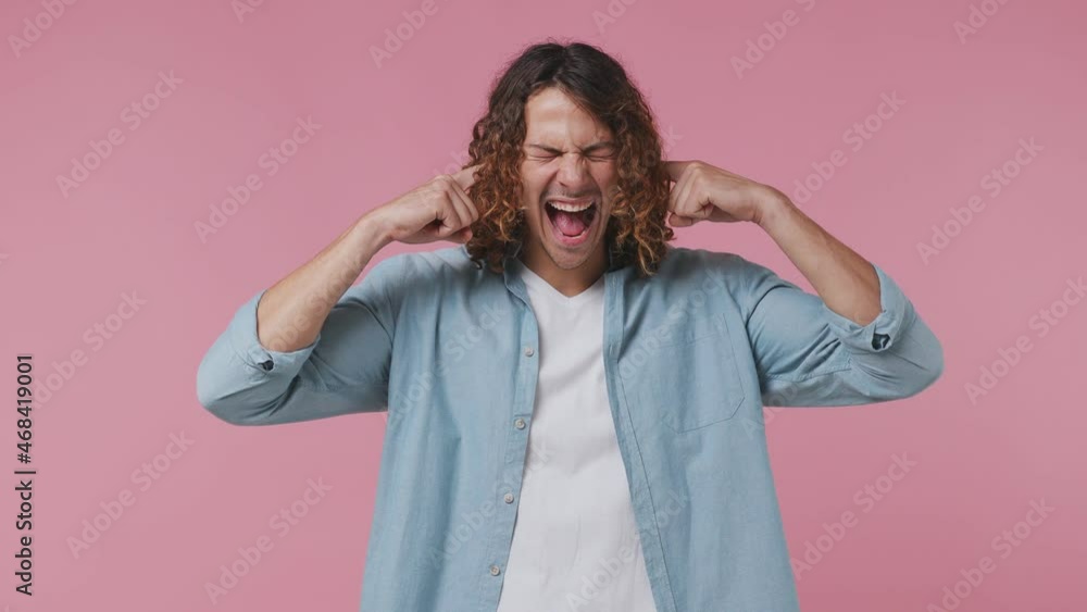 Displeased irritated nervous angry young curly man 20s wears blue shirt white t-shirt closed eyes cover ears do not want to listen scream isolated on pastel plain light pink background studio portrait Displeased irritated nervous angry young curly man 20s wears blue shirt white t-shirt closed eyes cover ears do not want to listen scream isolated on pastel plain light pink background studio portrait