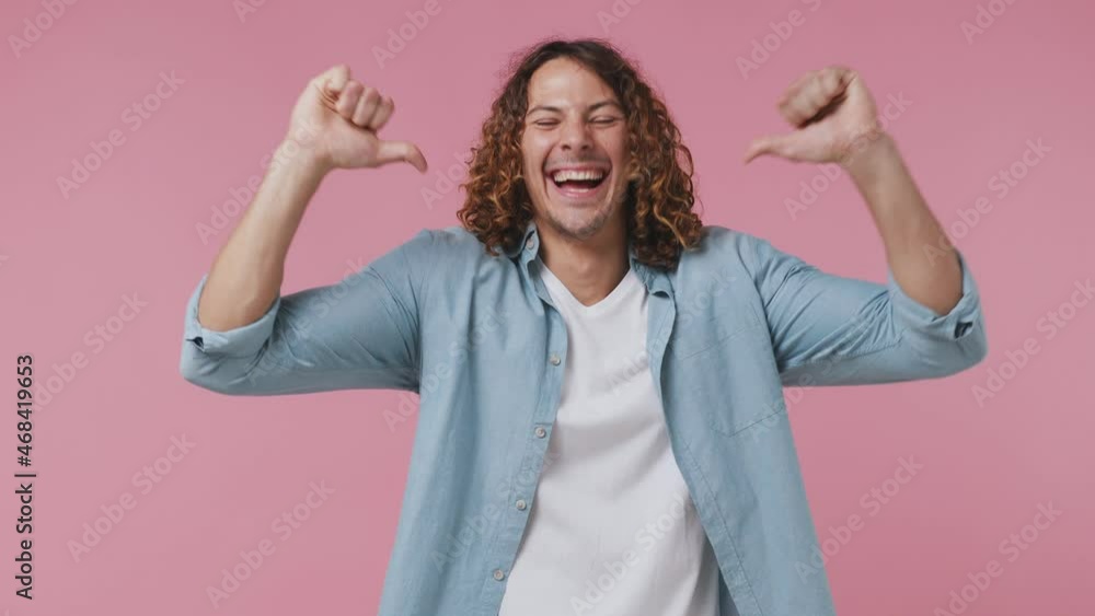 Confident lucky fun young curly man 20s wears blue shirt white t-shirt pointing fingers on himself blinking showing thumb up like gesture isolated on pastel plain light pink background studio portrait Confident lucky fun young curly man 20s wears blue shirt white t-shirt pointing fingers on himself blinking showing thumb up like gesture isolated on pastel plain light pink background studio portrait