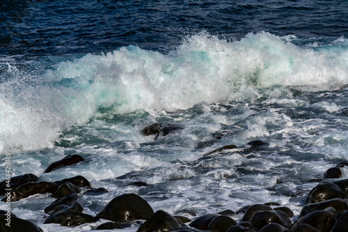 Breaking wave at volcanic pebble beach on the north coast of Madeira island Portugal near Porto Moniz. Spray, splashes and drops of turquoise blue sea water on a sunny day with perfect surf.