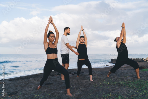 Caucasian yogi in casual sportswear teaching asanas male and female during together balance practice at coastline beach, group of concentrated persons training at seashore listening mentor advices