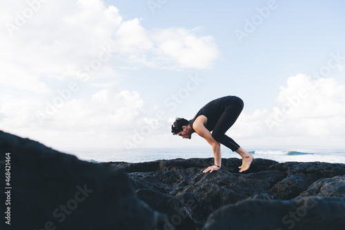 Energetic man in tracksuit doing balance Pose during yoga exercises in morning, Caucasian male yogi enjoying hatha practice on hands during time for physical recreation near coastline seashore