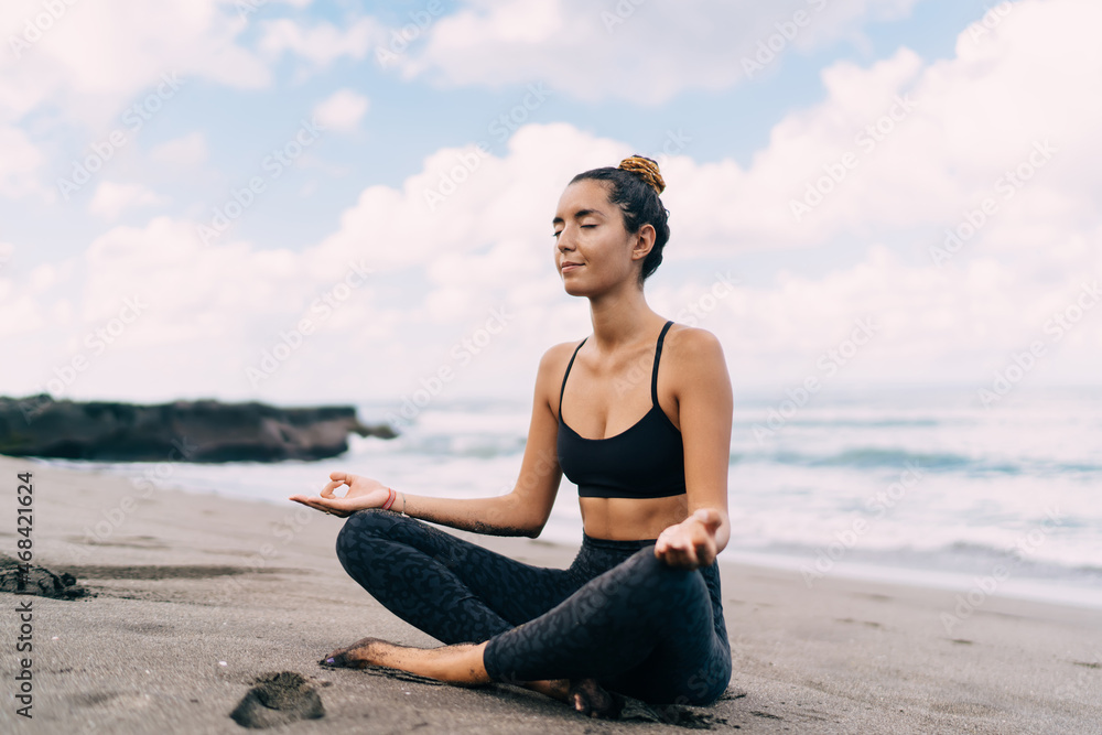 Middle Eastern female yogi meditating in lotus pose getting energy from ocean seeking enlightenment and getting mindfulness harmony, calm woman relaxing during concentration workout at coastline