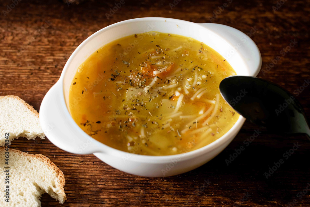 Chicken soup with noodles, potatoes, onions and carrots in a white deep plate with a black spoon and white bread on a wooden background. Chicken soup with noodles, potatoes, onions and carrots in a white deep plate with a black spoon and white bread on a wooden background.