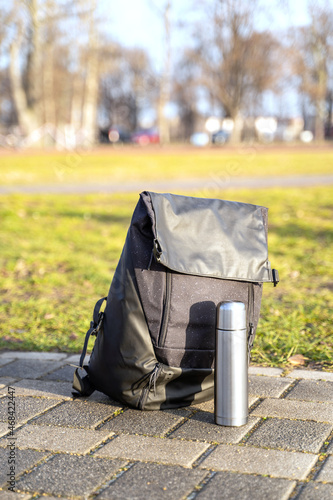 Sports waterproof fabric modern stylish black large backpack lies next to metal thermos on brick path in park or sports ground in early morning at sunrise or sunset on autumn, winter or spring day