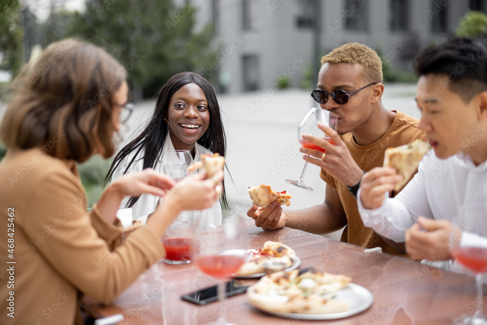 Multiracial business team having lunch with wine and pizza at outdoor cafe. Concept of teambuilding and corporate event. Idea of rest and leisure on job. People sitting at wooden table and talking
