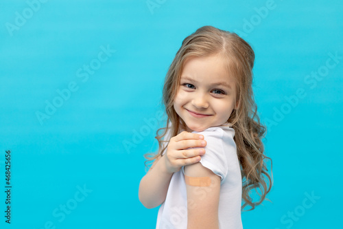 a little girl child with a patch on her arm after vaccination in a white T-shirt on a blue background in the studio, space for text, the concept of vaccination, health and medicine