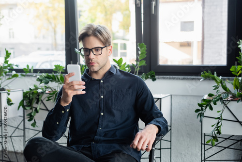 Young developer in eyeglasses using smartphone while sitting on chair in office