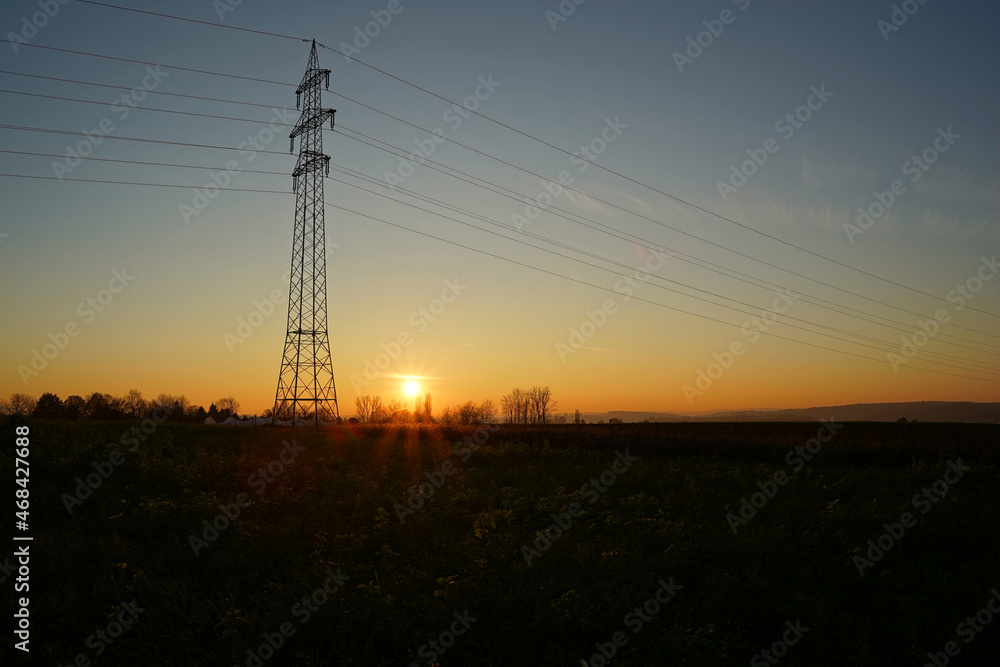 Single Electricity pylon (Strommasten) also overhead line pylon on the field. The sun sets on a clear evening sky. Side View.
