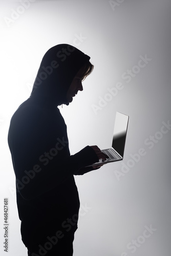 Side view of hacker in hood using laptop isolated on grey
