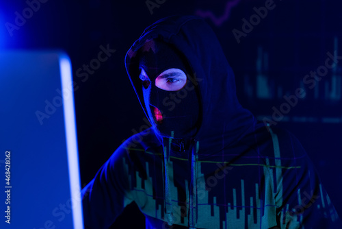 Hacker in balaclava with charts reflection looking at blurred computer monitor on black background