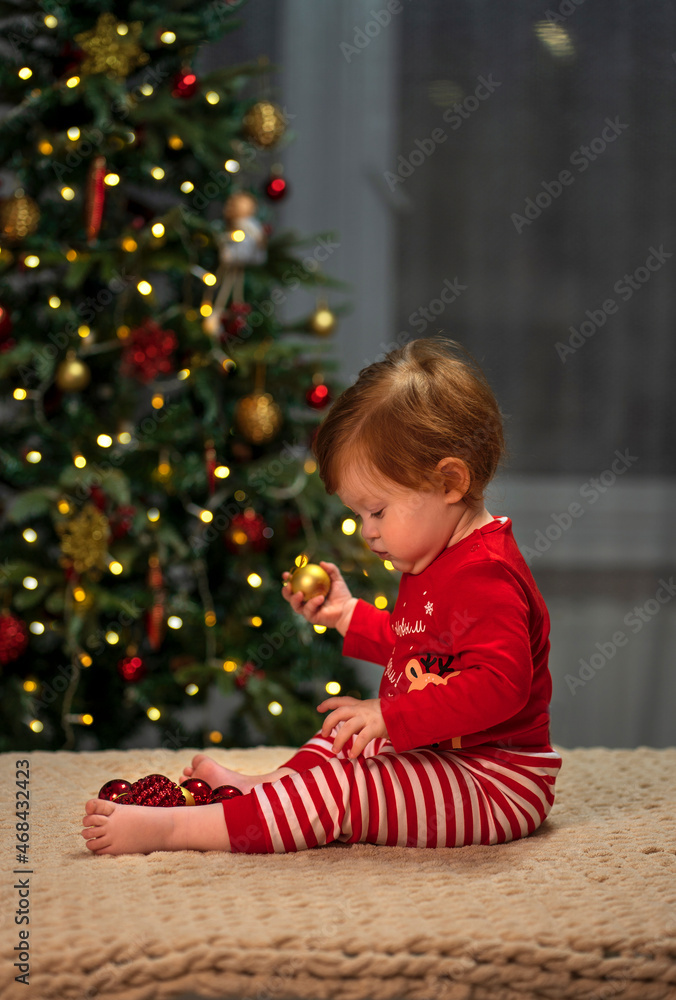 A small child with red hair and a red Christmas costume sits at home against the background of a Christmas tree and plays with Christmas decorations A small child with red hair and a red Christmas costume sits at home against the background of a Christmas tree and plays with Christmas decorations