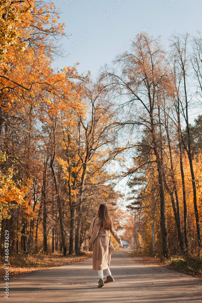 Young beautiful woman in beige coat walking on the road in autumn park