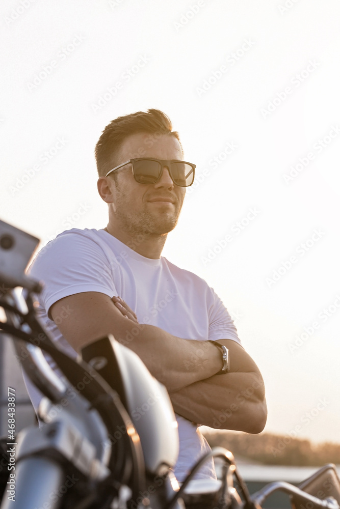Portrait of a young attractive guy in sunglasses. Nice young man in a white T-shirt and black jeans on a motorcycle. Life style photography. Summer evening sky background. Vertical shot