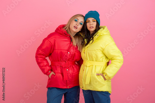 two attractive stylish women posing on pink background in colorful winter down jacket of red and yellow color, warm clothes fashion trend
