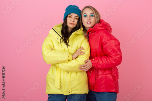 two attractive stylish women posing on pink background in colorful winter down jacket of red and yellow color, warm clothes fashion trend