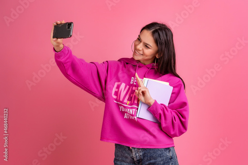 hispanic pretty woman in pink hoodie smiling holding holding notebooks and using smartphone