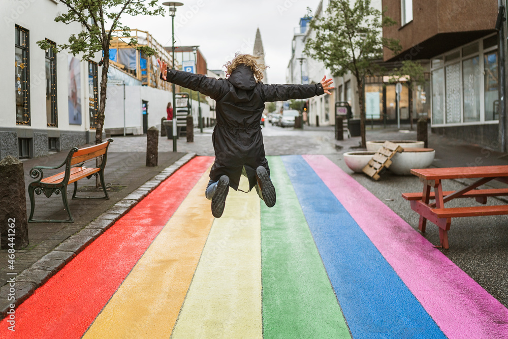 Anonymous boy jumping for joy in the streets of Reykjavik, Iceland on a roadway painted in the colours of the LGTBI+ rainbow flag. Celebrating sexual equality. - obrazy, fototapety, plakaty 