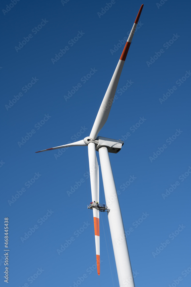 Red and white wind turbine for power generation with a maintenance team working in a basket on one blade with a blue sky in the background
