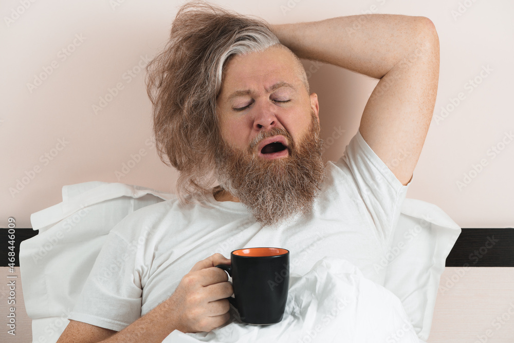 Yawning bearded man drinking morning coffee lying in bed. Sleepy guy trying to wake up. Closeup.