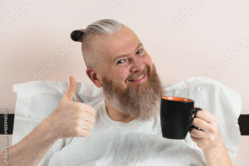 Happy bearded man enjoying morning coffee in bed showing thumb up gesture. Smiling hipster guy starting the day. Closeup.
