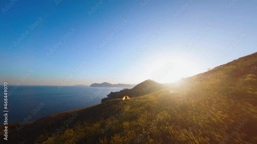 Speed fpv drone flying through tent in mountain on seashore. Dynamic low motion above hil slope covered grass and happy woman tourist standing on cliff with rising hands at sunset time Speed fpv drone flying through tent in mountain on seashore. Dynamic low motion above hil slope covered grass and happy woman tourist standing on cliff with rising hands at sunset time