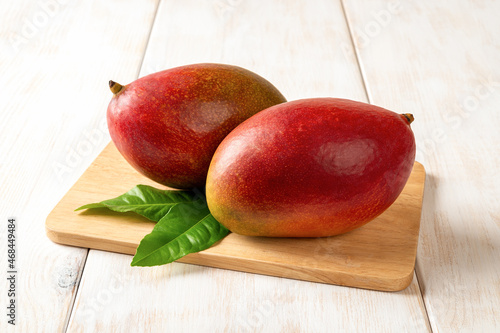 Two red ripe Palmer mangoes on a cutting board over white wooden table. Ready to eat whole sweet tasty mango. Two delicious tropical fruits of Mangifera Indica close-up. photo